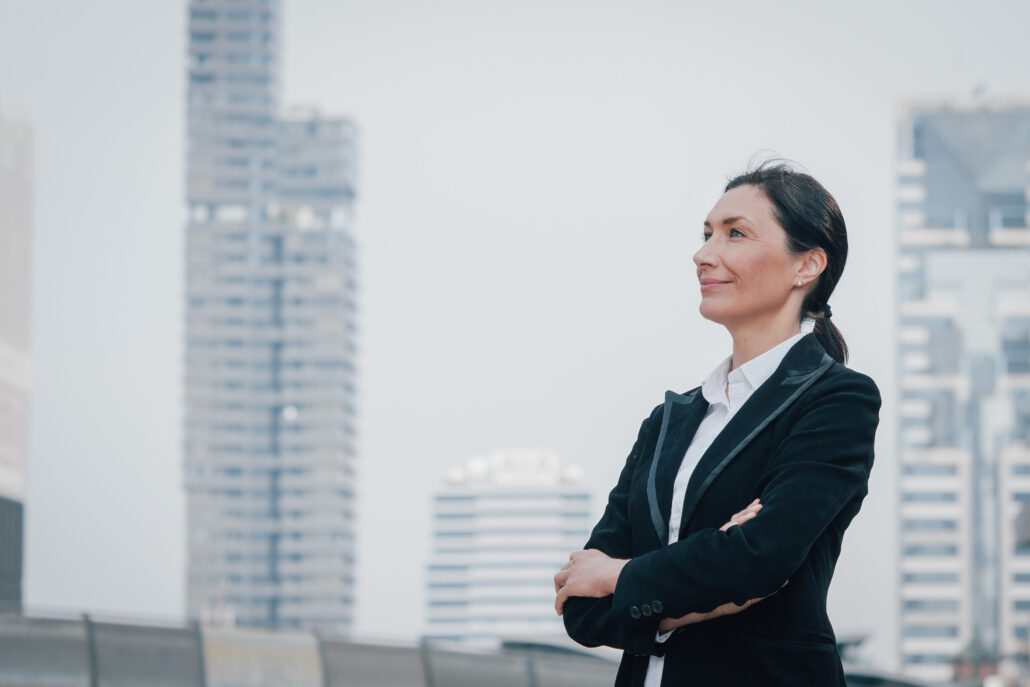 A woman in a suit stands confidently with arms crossed, looking to the side. She is outdoors in a cityscape with tall buildings in the background.